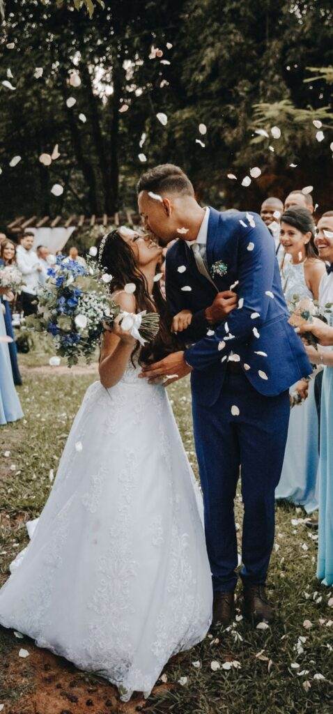 A newlywed couple kisses outdoors under a shower of white rose petals. The bride, in a white gown, holds a bouquet of blue and white flowers. The groom, in a blue suit, embraces her. Guests, dressed in blue, smile in the background amidst green foliage.