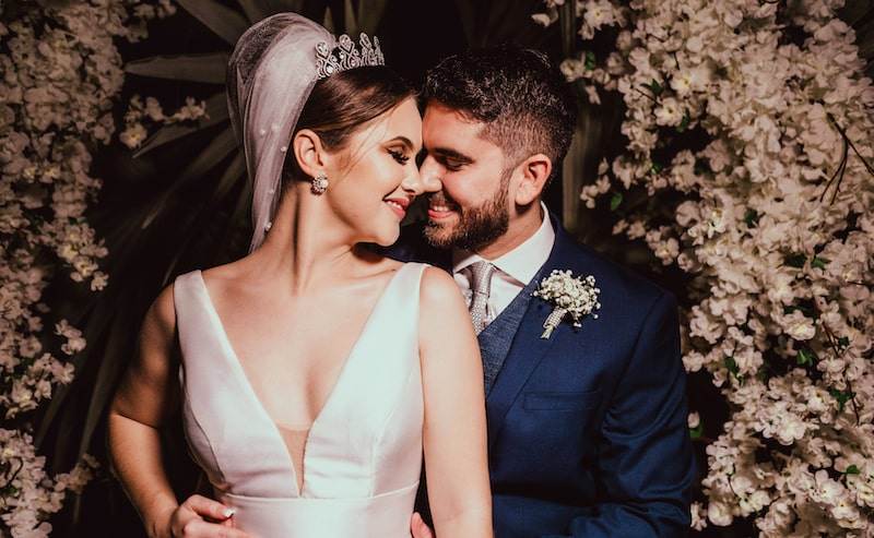 A bride and groom stand close together, smiling joyfully. The bride wears a white dress and veil, and the groom is in a navy blue suit with a white shirt and tie. They are surrounded by an arch of white flowers.