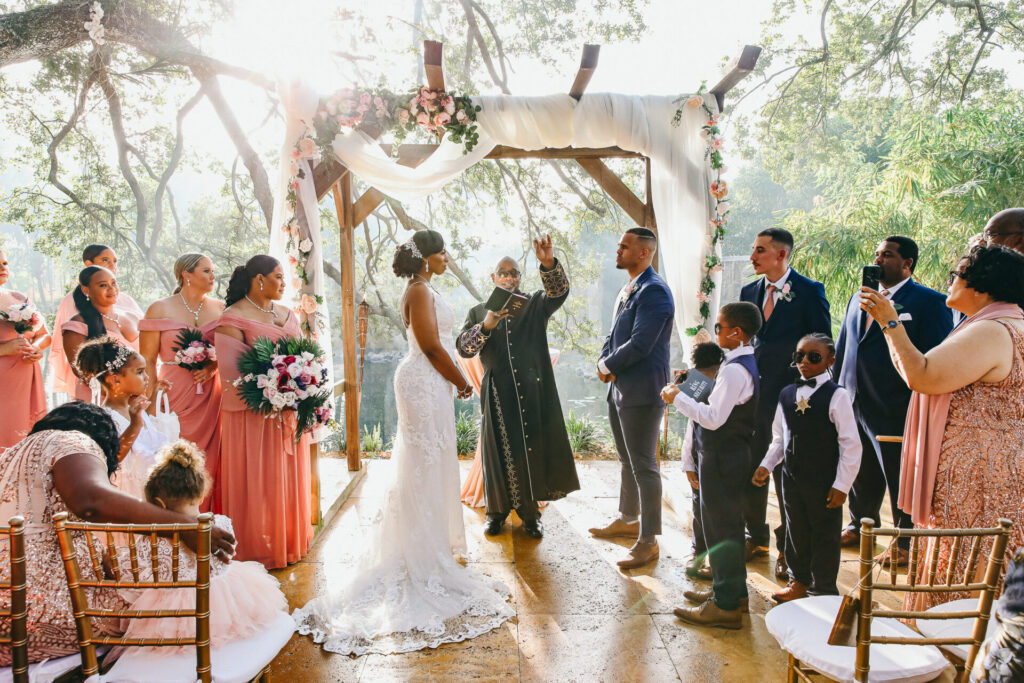 Bride and groom standing under a floral arch during an outdoor wedding ceremony with bridesmaids and groomsmen looking on.