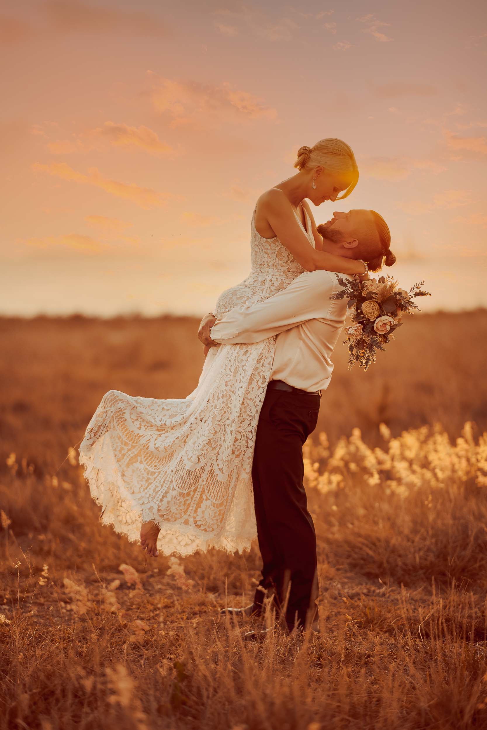 A romantic wedding scene at sunset, with the groom lifting the bride in a golden field, surrounded by the warm glow of the evening light.